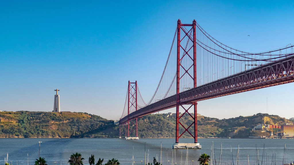 A scenic view of the 25 de Abril Bridge in Lisbon, Portugal, with its striking red design spanning across the Tagus River. In the background stands the Cristo Rei statue on a hill, surrounded by greenery and clear blue skies.