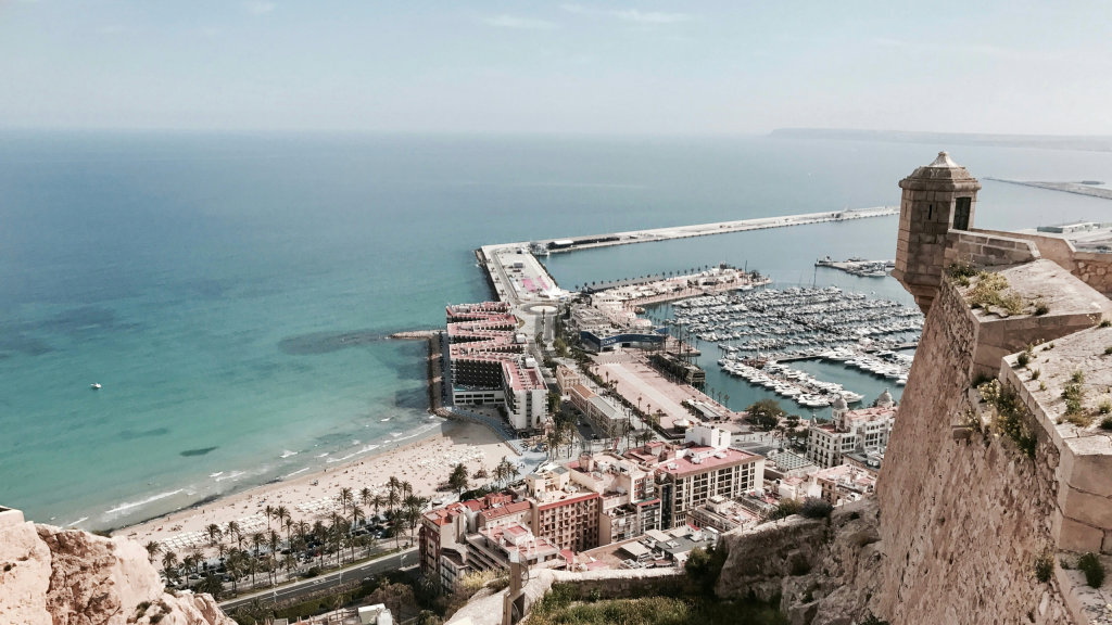 A panoramic view of a coastal city featuring a marina with numerous yachts, a sandy beach lined with palm trees, and historical architecture on a cliffside overlooking the sea.